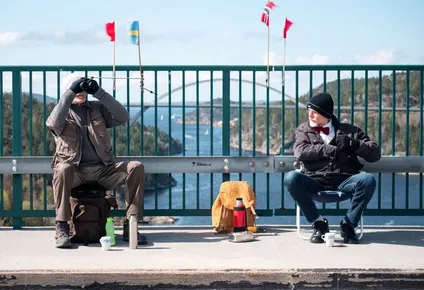 Pontus Berglund (L) looks at birds into his binoculars as he sits on the Swedish side while his brother Ola sits on the Norwegian side of the old bridge of Svinesund and the new Svinesund Bridge is seen in the background, in Svinesund, Norway and Sweden, on May 1, 2021. They havent been allowed to have each other over for a year because of Covid. So come rain or shine, two 73-year-old Swedish twins have met every Saturday, each on their side of the border, on a bridge that links Norway and Sweden.
